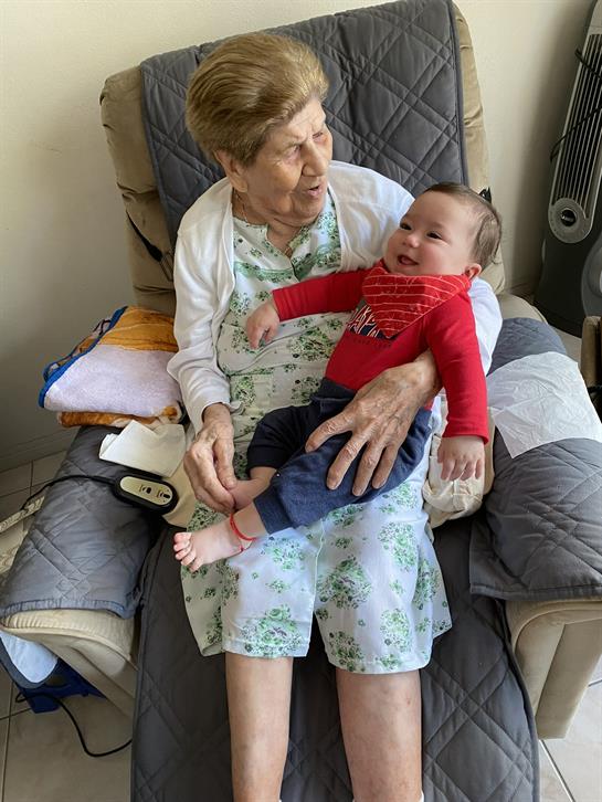 An elderly woman smiles while holding a baby on her lap in a cozy living room setting.