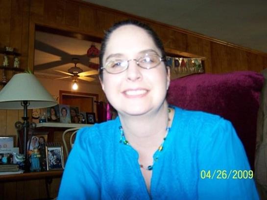 Woman with glasses smiles warmly while sitting in a cozy living room decorated with family photos.