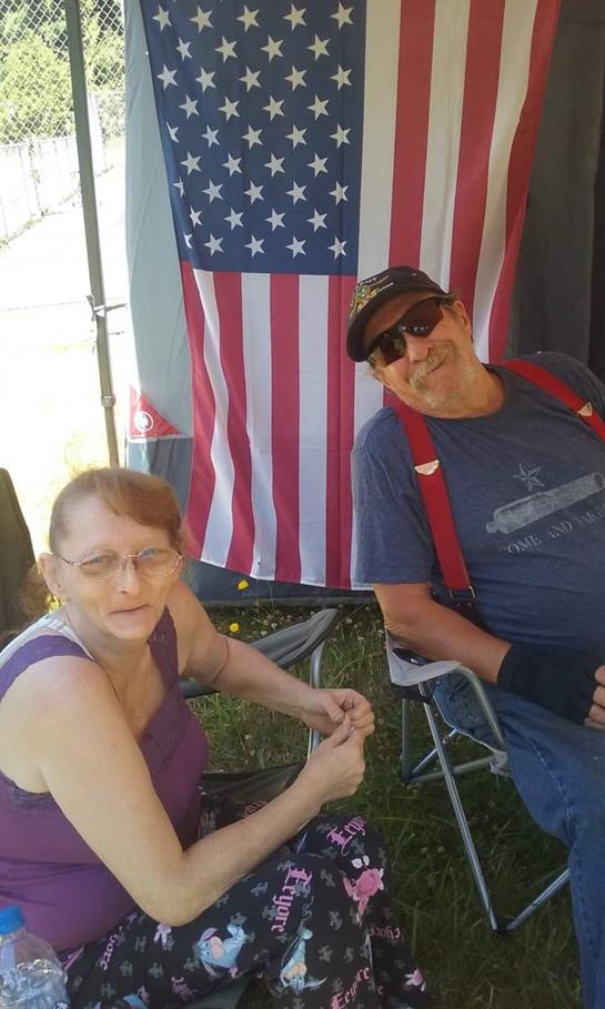 Two individuals sit in folding chairs near an American flag, chatting and enjoying the weather.