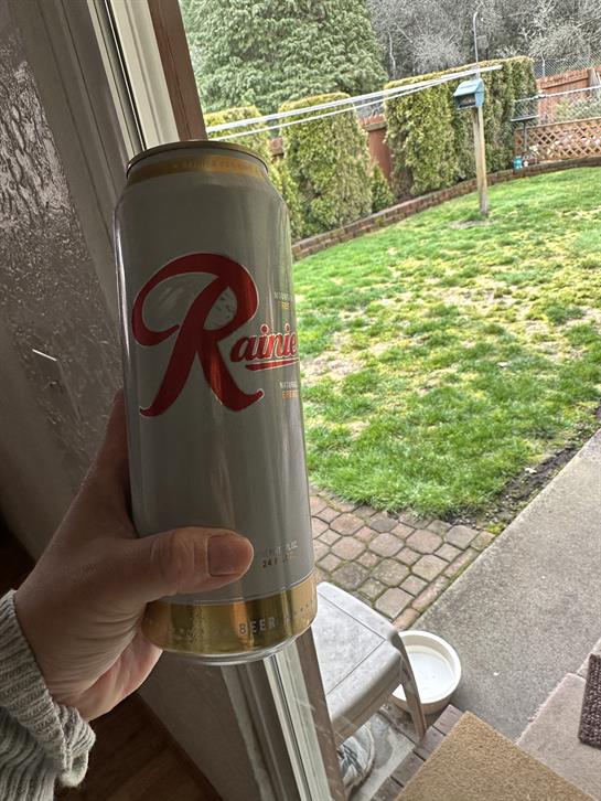 A person holds a tall can of beverage while standing near an open door to a backyard.