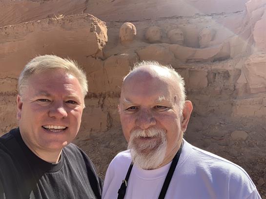 Two men enjoy time together, smiling at the camera with a natural rock formation behind them.