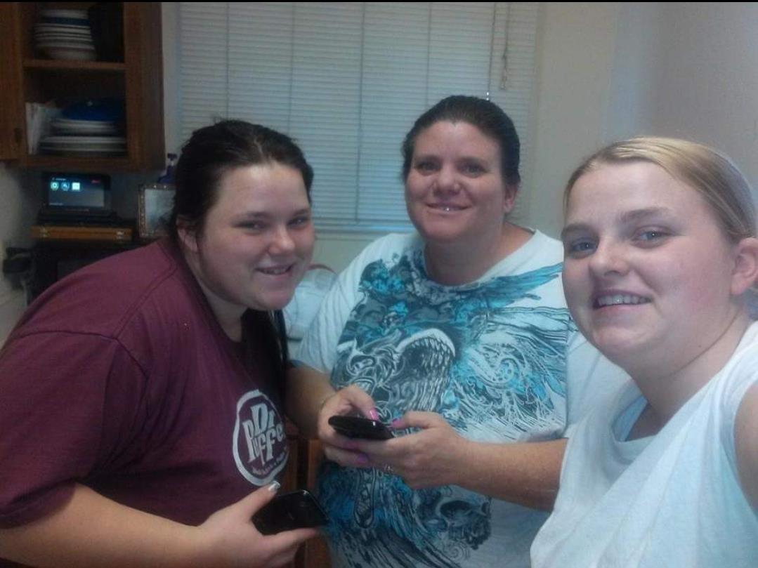 Three friends smile while interacting with their phones in a warm kitchen at night.
