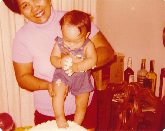 A joyful woman holds a toddler on a cake during a birthday celebration, capturing the festive mood.