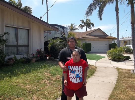 Two people pose for a casual picture outside a house in a sunny suburban environment.
