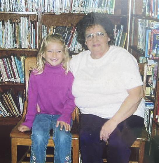 A child and an older woman sit side by side in a cozy library full of bookshelves.