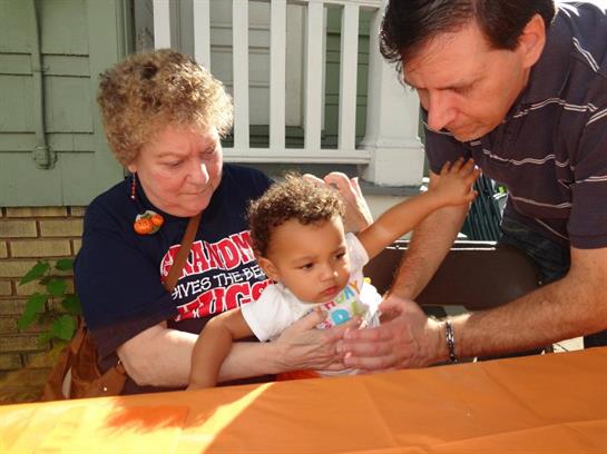 A caregiver helps a baby reach for a toy while another family member observes joyfully.