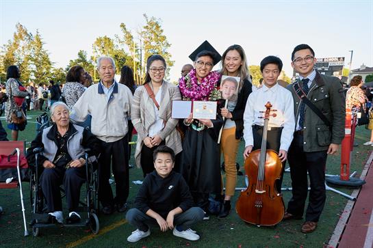 A proud graduate stands with family and friends holding a diploma, celebrating achievement outdoors.