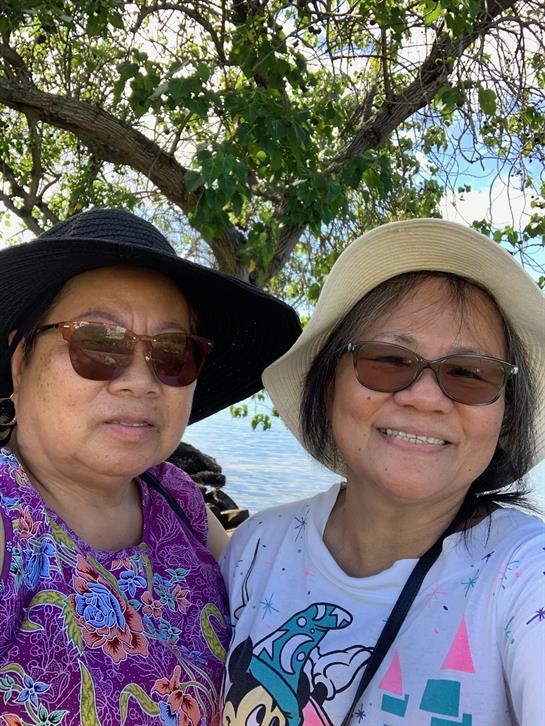 Two women smile joyfully as they pose under a tree near the beach on a sunny day.