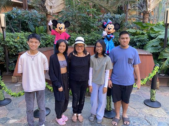 Group of five people poses happily at a tropical resort in front of Disney characters outdoors.