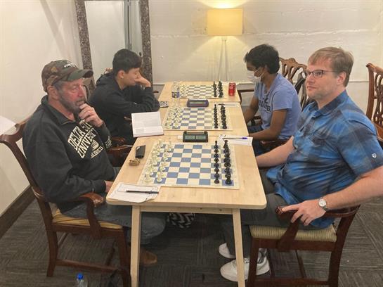 Four people engage in a friendly chess game at a small table in a casual setting.