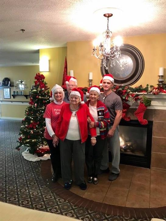 Five people in festive attire gather by a Christmas tree and fireplace in a community space.