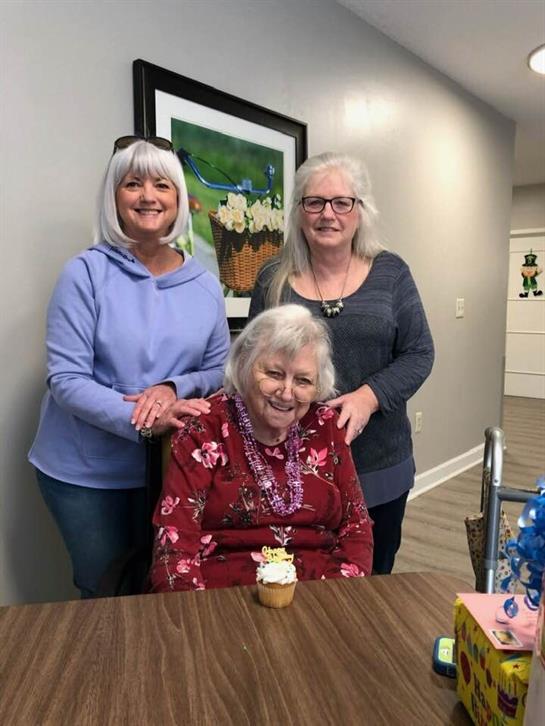 Three women gather around a birthday cupcake, smiling joyfully in a welcoming indoor space.