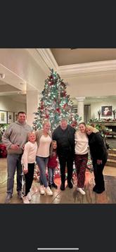 Family members happily pose by a tall, festive Christmas tree in a cozy living room.