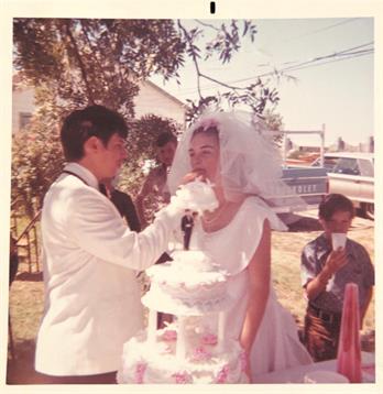 A joyful couple celebrates their wedding with a cake cutting ceremony in a sunny outdoor setting.
