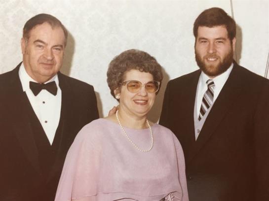 Two men in tuxedos and a woman in a purple dress stand together, smiling proudly at an event.