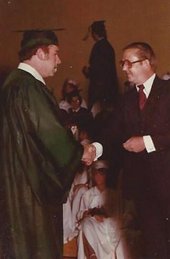 Two men shake hands at graduation, while others celebrate this educational milestone.