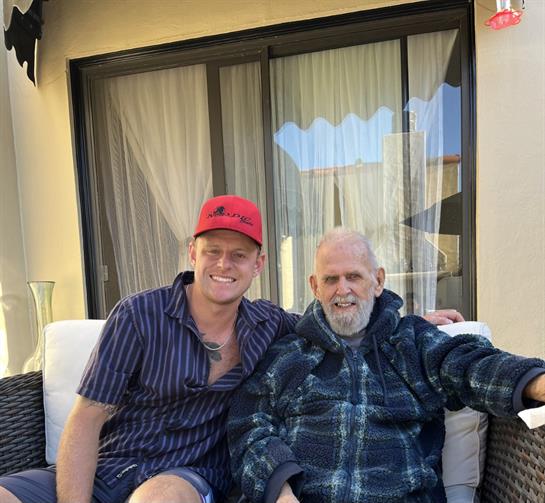 Two men share a joyful moment sitting together outdoors, enjoying a sunny day by the house.