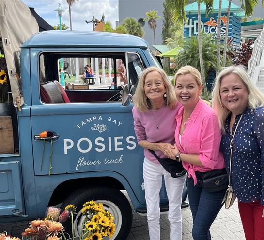 Three friends smile joyfully as they stand near a flower truck at a lively Tampa market.
