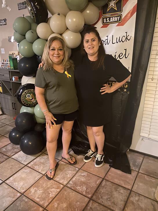 Two women smile and stand together in a celebration setting with colorful balloons and decor.
