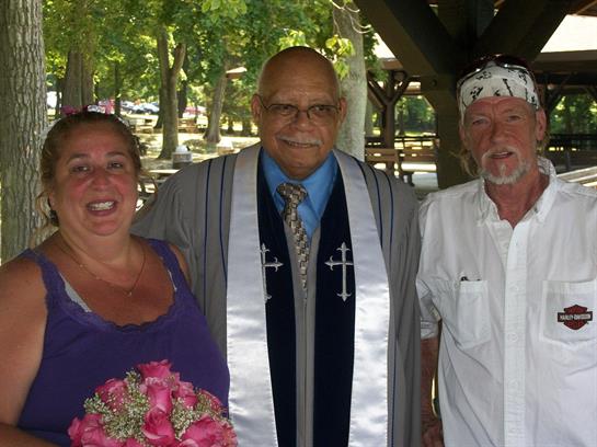 A couple smiles with their officiant, celebrating their wedding in a sunny park.