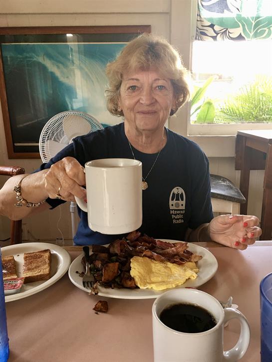 A cheerful woman holds a large cup of coffee while sitting at a table filled with breakfast food.