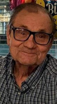 Elderly man with glasses and a straw hat smiles warmly while seated indoors next to a window.