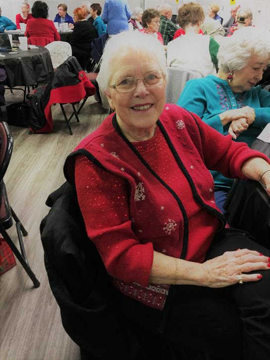 A cheerful elderly woman in a red sweater enjoys a social gathering, surrounded by friends.