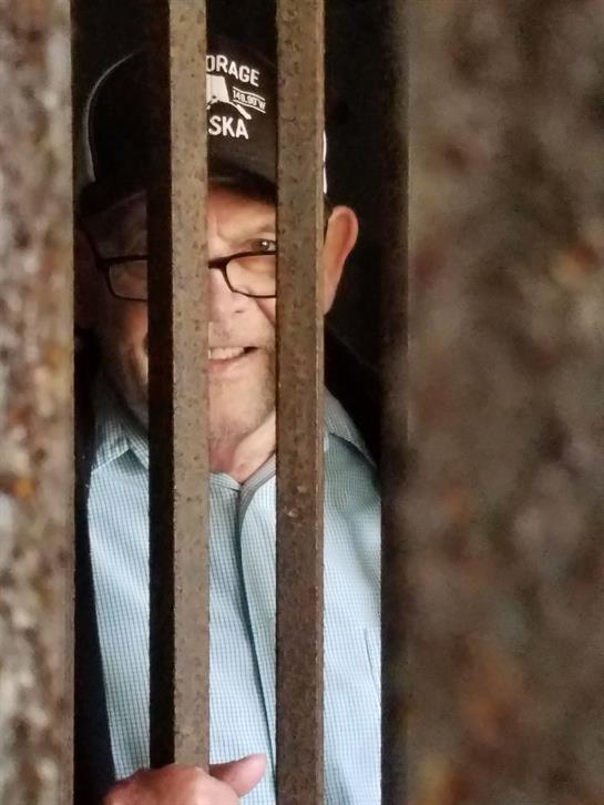 An old man with glasses peers through rusty bars, curiosity evident in an urban setting.