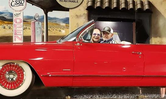 An elderly couple smiles joyfully from a classic red convertible parked at a retro gas station.