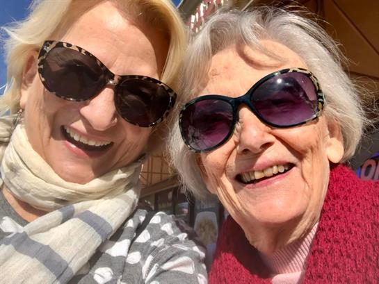 Two joyful women share a moment of laughter in a bustling outdoor area under bright sunlight.