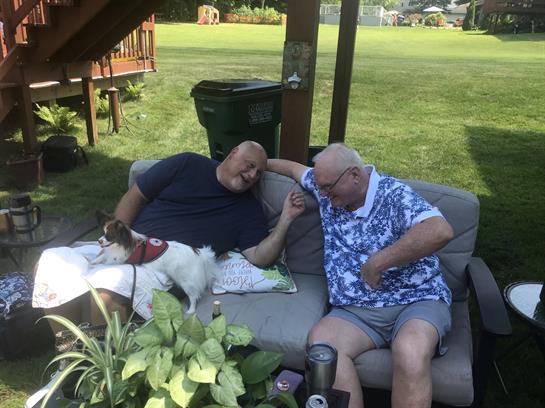 Two men sit comfortably on a couch, enjoying each other's company in a sunny outdoor setting.