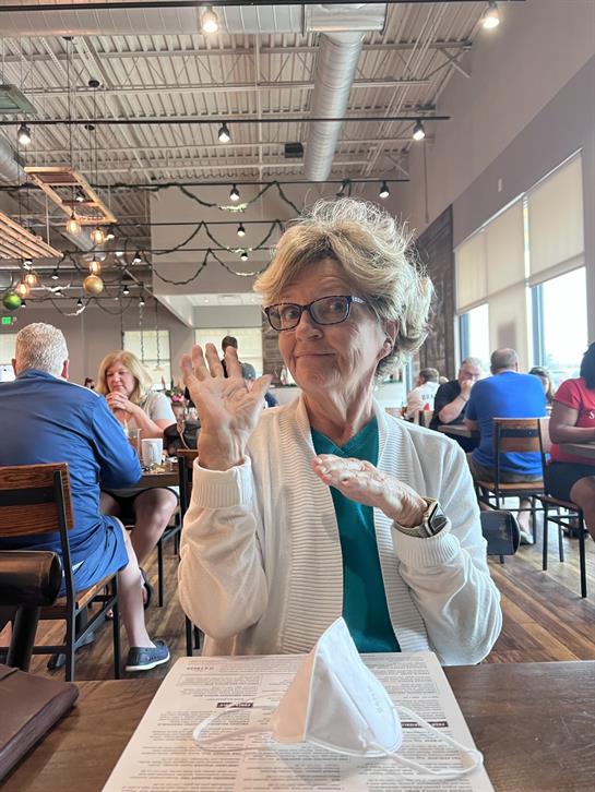 A woman expresses excitement with hand gestures while sitting at a table in a crowded restaurant.