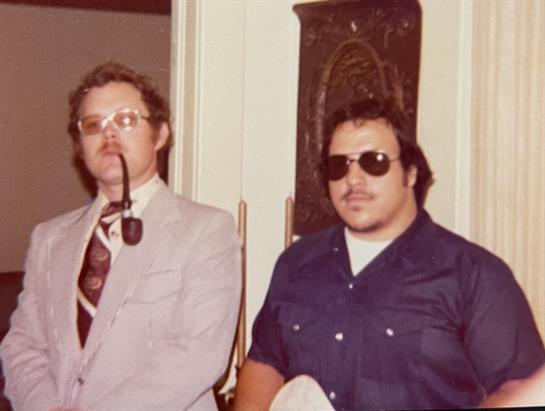 Two men stand side by side, one wearing glasses and a pipe, and the other in sunglasses, smiling.