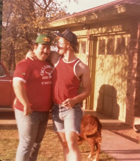 Two friends pose playfully outdoors near a vintage vehicle and a brown dog on a sunny day.