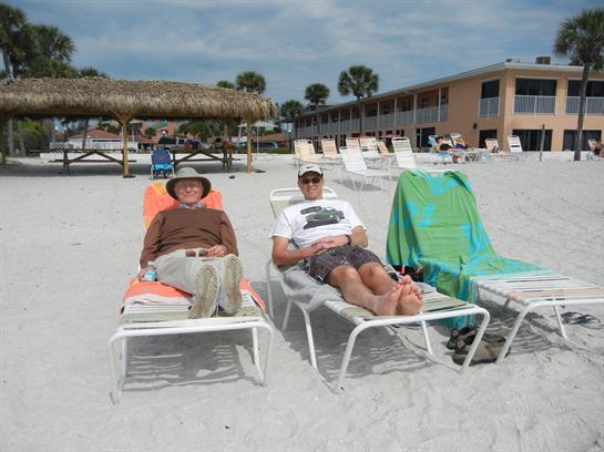Two friends lounge on beach chairs, soaking up the sun near a resort on a sandy shore.