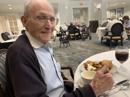 Elderly gentleman smiles while holding a plate of food in a cozy dining area.
