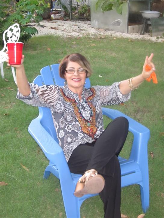 Woman relaxes in blue chair, holding a drink and enjoying a cheerful moment outdoors.
