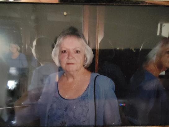 An elderly woman with white hair stands in an indoor setting, looking directly at the camera.