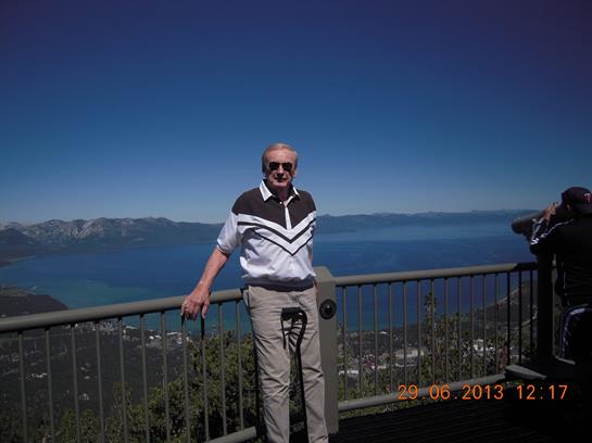 An elderly man stands at a lookout point, admiring the stunning view of a lake and mountains.