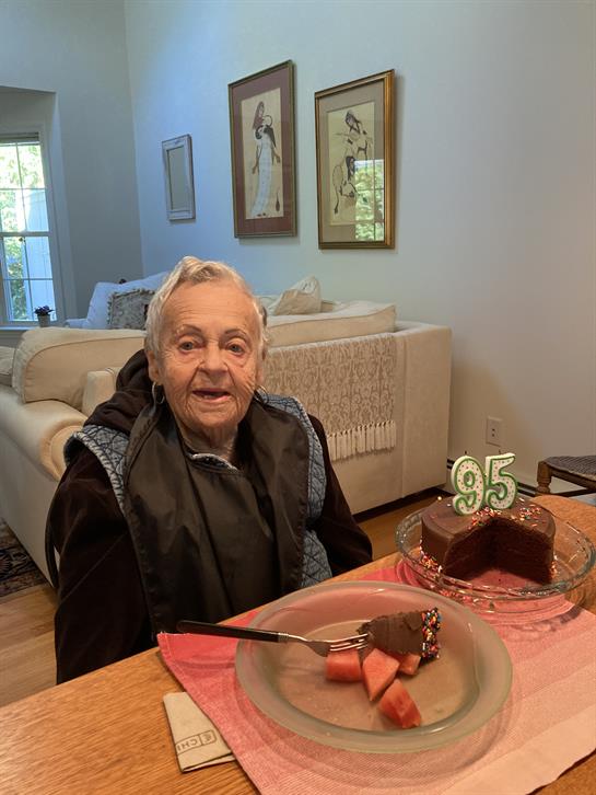 A man enjoys his 95th birthday celebration at home, sitting with a chocolate cake and smiles.
