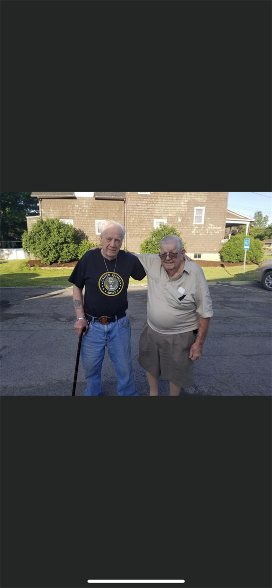 Two older gentlemen stand together outdoors, smiling and enjoying each other's company.