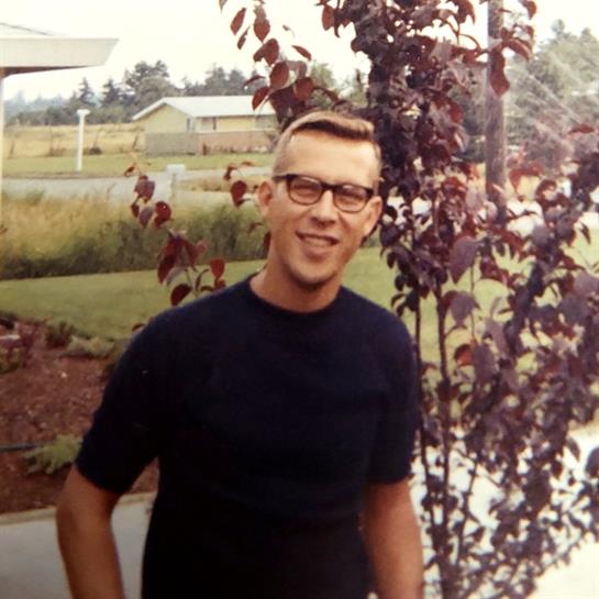A young man poses and smiles in a yard surrounded by trees and greenery on a sunny day.