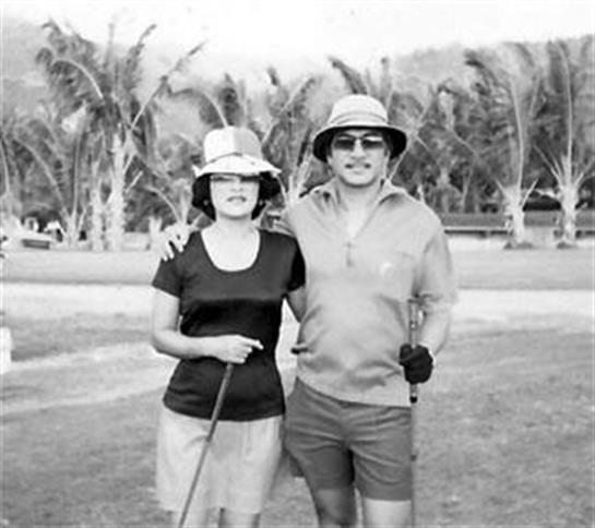 A couple poses with golf clubs on a sunny day at a tropical resort surrounded by palm trees.