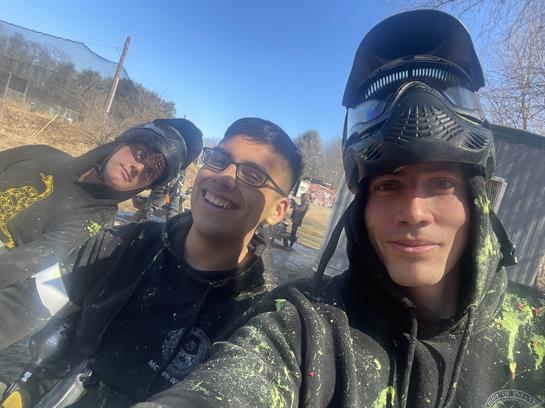 Three young men are smiling and posing while experiencing paintball in an outdoor arena.