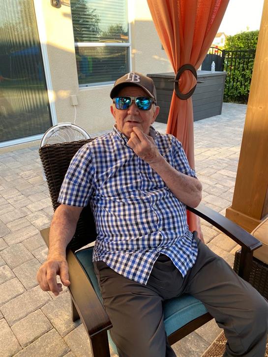 A man wearing sunglasses and a checkered shirt sits thoughtfully in a patio chair.