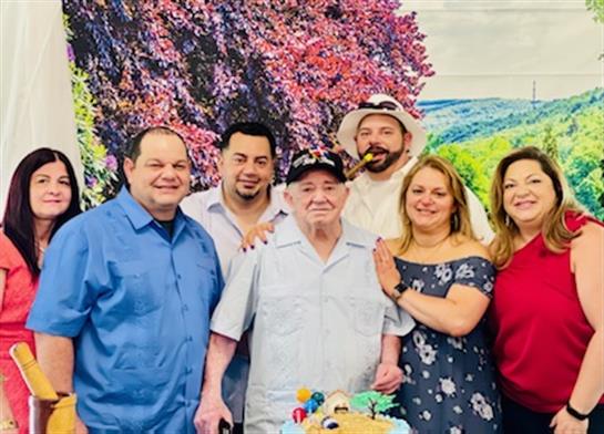 A group of six people joyfully poses together with a colorful cake in a lively environment.