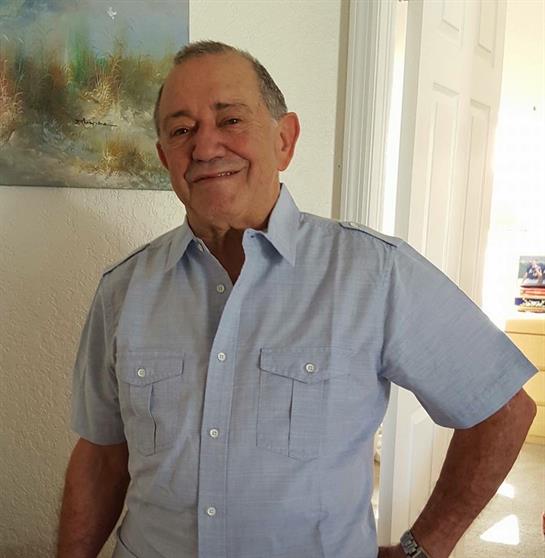 An elderly man in a blue shirt smiles warmly while standing in a well-lit room decorated with art.