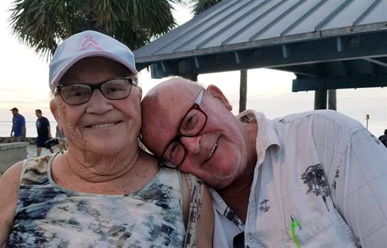 Two smiling elderly individuals share a joyful embrace, surrounded by palm trees at sunset.