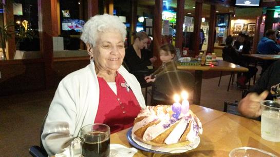 A joyful elderly woman enjoys her birthday cake, surrounded by family in a bustling restaurant.