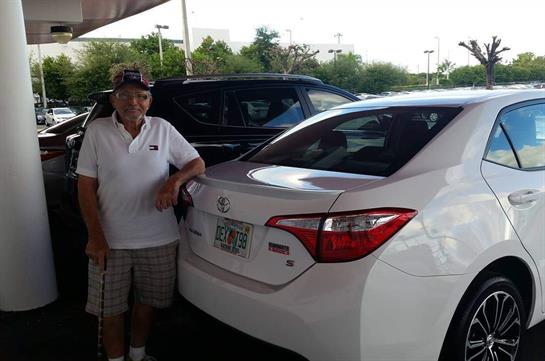 Smiling elderly couple stands beside their car in a sunny shopping complex parking lot.